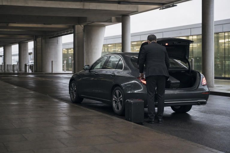 A driver lifts a suitcase into the open boot of a dark saloon at an airport terminal drop-off kerb.