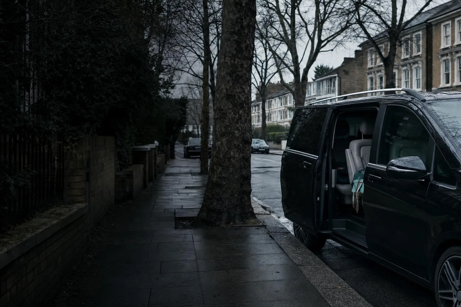 A dark people-carrier with its sliding door open, parked at the kerb of a quiet suburban street beside a low brick wall.