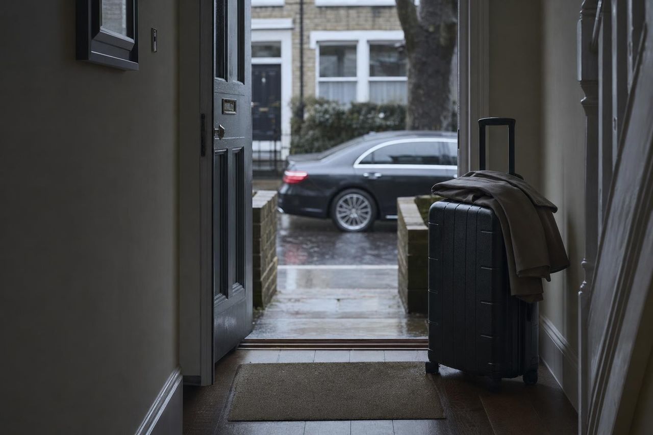 A suitcase and folded coat waiting in a hallway at dawn, the front door ajar onto a damp street with a car waiting beyond.